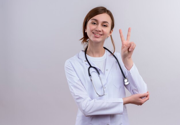 Sorridente giovane medico femminile che indossa abito medico e stetoscopio facendo segno di pace sulla parete bianca isolata con lo spazio della copia