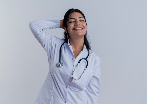 Sorridente giovane medico femminile che indossa abito medico e stetoscopio mettendo la mano dietro la testa con gli occhi chiusi, isolato sulla parete bianca con lo spazio della copia