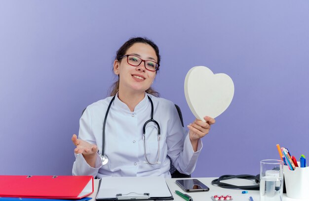 Sorridente giovane medico femminile che indossa abito medico e stetoscopio seduto alla scrivania con strumenti medici che tengono la forma del cuore che mostra la mano vuota che sembra isolata