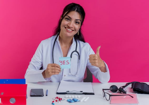 Sorridente giovane medico femminile che indossa abito medico e stetoscopio seduto alla scrivania con strumenti medici che tengono la nota di sì che mostra il pollice in su isolato sulla parete rosa