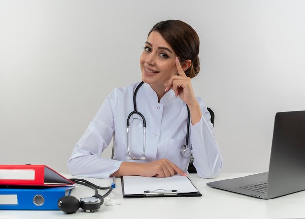 Sorridente giovane medico femminile che indossa abito medico e stetoscopio seduto alla scrivania con strumenti medici e computer portatile toccando la testa con il dito isolato sul muro bianco