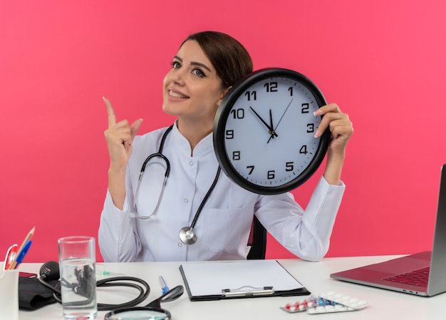 Sorridente giovane medico femminile che indossa abito medico e stetoscopio seduto alla scrivania con strumenti medici e laptop tenendo l'orologio alla ricerca e rivolto verso l'alto isolato sul muro rosa