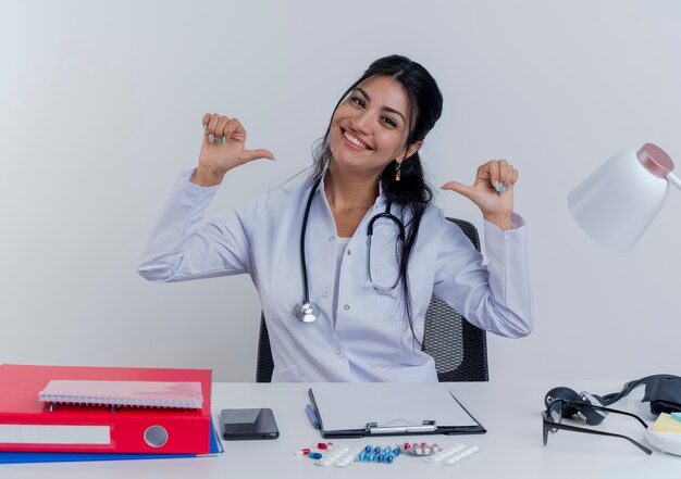 Sorridente giovane medico femminile che indossa abito medico e stetoscopio seduto alla scrivania con strumenti medici guardando che punta a se stessa isolato