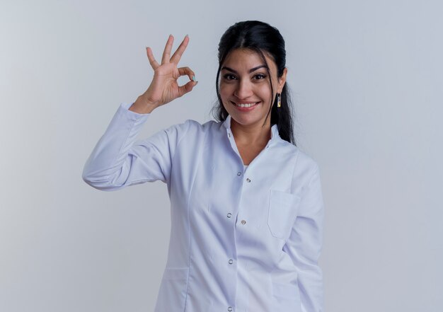 Sorridente giovane medico femminile che indossa abito medico facendo segno ok guardando isolato