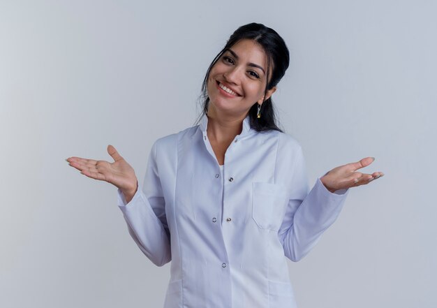 Sorridente giovane medico femminile che indossa abito medico guardando mostrando le mani vuote isolate