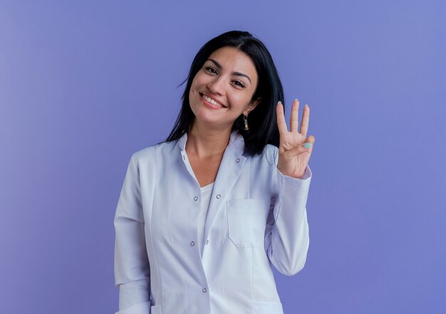 Sorridente giovane medico femminile che indossa abito medico guardando mostrando tre con mano