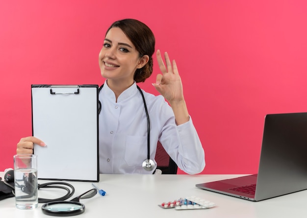 Sorridente giovane medico femminile che indossa veste medica e stetoscopio seduto alla scrivania con strumenti medici e computer portatile che mostra appunti e facendo segno ok isolato sul muro rosa