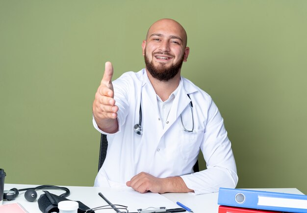 Sorridente giovane medico maschio calvo che indossa abito medico e stetoscopio seduto al lavoro di scrivania