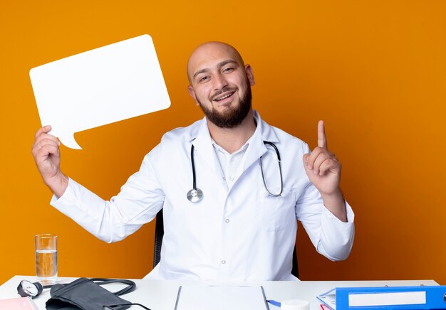 Sorridente giovane medico maschio calvo che indossa abito medico e stetoscopio seduto alla scrivania con strumenti medici che tengono bolla di chat e punti in alto isolato sulla parete arancione