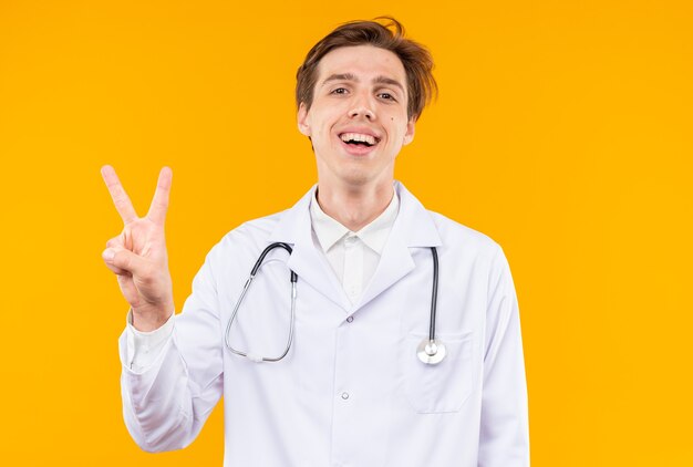 Sorridente giovane medico maschio che indossa abito medico con stetoscopio che mostra gesto di pace isolato su parete arancione