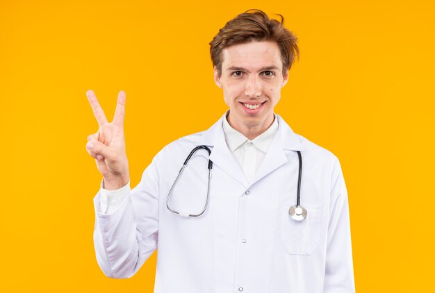 Sorridente giovane medico maschio che indossa abito medico con stetoscopio che mostra gesto di pace