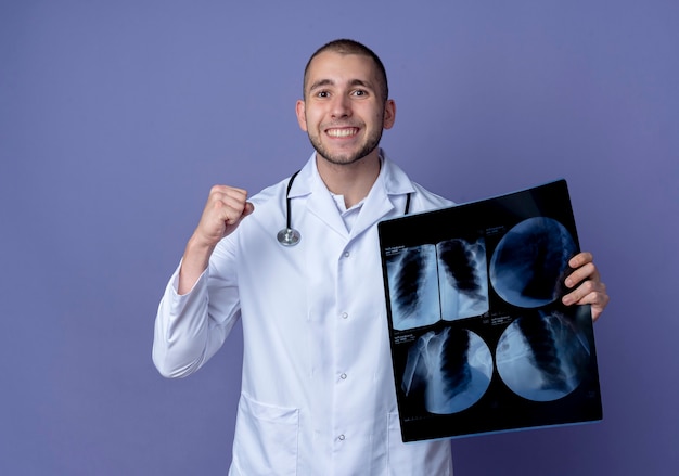 Sorridente giovane medico maschio che indossa abito medico e stetoscopio che tiene il colpo dei raggi x e il pugno di serraggio isolato sulla parete viola