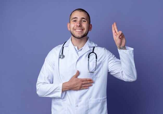 Sorridente giovane medico maschio che indossa abito medico e stetoscopio facendo gesto di promessa isolato sulla parete viola