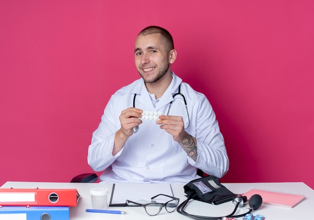 Sorridente giovane medico maschio che indossa abito medico e stetoscopio seduto alla scrivania con strumenti di lavoro che tengono il pacchetto di compresse isolato sulla parete rosa