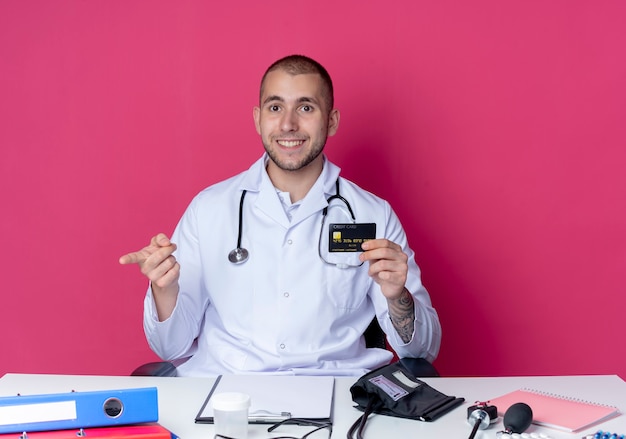 Sorridente giovane medico maschio che indossa abito medico e stetoscopio seduto alla scrivania con strumenti di lavoro in possesso di carta di credito che punta dritto isolato sulla parete rosa