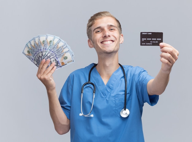 Sorridente giovane medico maschio che indossa l'uniforme del medico con lo stetoscopio che dà contanti e carta di credito alla macchina fotografica isolata sul muro bianco