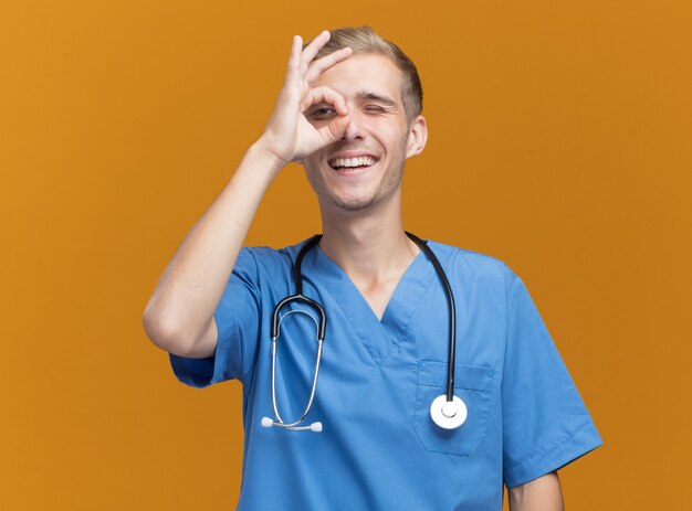 Sorridente giovane medico maschio che indossa l'uniforme del medico con lo stetoscopio che mostra il gesto di sguardo isolato sulla parete arancione