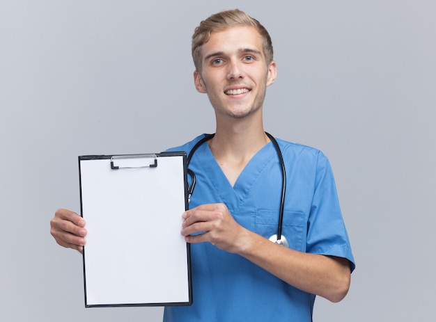 Sorridente giovane medico maschio che indossa l'uniforme del medico con lo stetoscopio che tiene appunti isolato sulla parete bianca