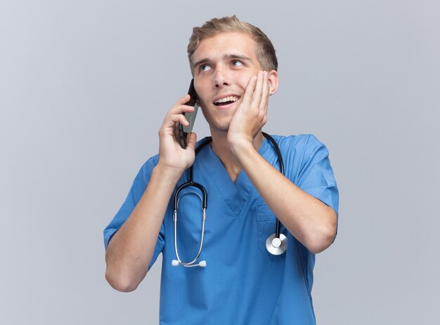 Sorridente giovane medico maschio che indossa l'uniforme del medico con lo stetoscopio parla al telefono mettendo la mano sulla guancia isolata sulla parete bianca