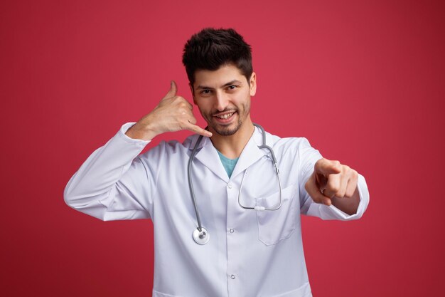 Sorridente giovane medico maschio che indossa uniforme medica e stetoscopio intorno al collo guardando e indicando la telecamera che mostra chiamami gesto isolato su sfondo rosso