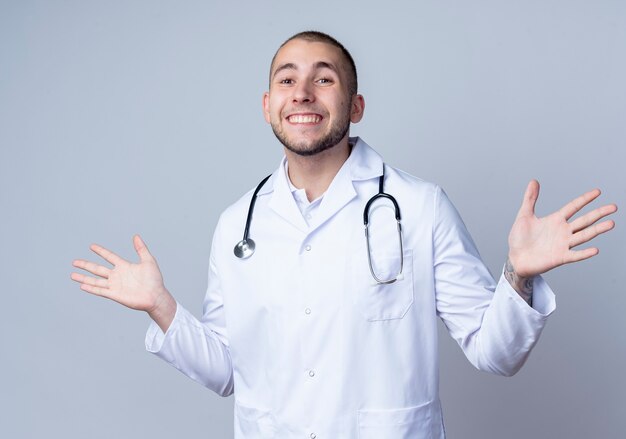 Sorridente giovane medico maschio che indossa veste medica e stetoscopio intorno al collo che mostra le mani vuote isolate sul muro bianco