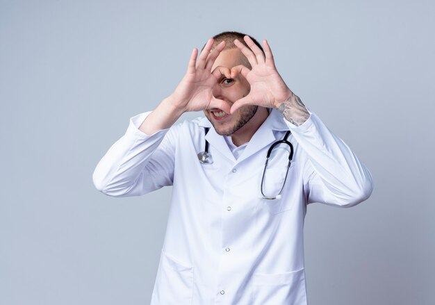 Sorridente giovane medico maschio che indossa veste medica e stetoscopio intorno al collo facendo il segno del cuore e guardando davanti attraverso di esso isolato sul muro bianco