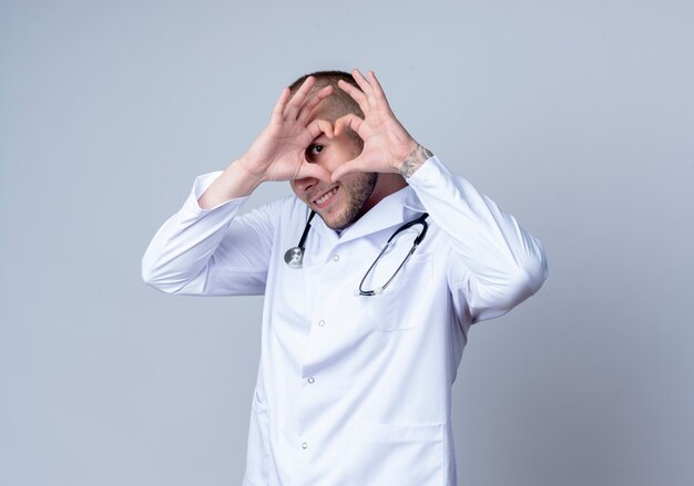 Sorridente giovane medico maschio che indossa veste medica e stetoscopio intorno al collo facendo il segno del cuore e guardando davanti attraverso di esso isolato sul muro bianco