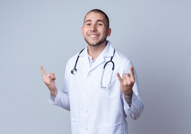 Sorridente giovane medico maschio che indossa veste medica e stetoscopio intorno al collo facendo segni di roccia isolato sul muro bianco