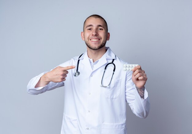 Sorridente giovane medico maschio che indossa veste medica e stetoscopio intorno al collo tenendo e indicando la confezione di compresse medicali isolato sulla parete bianca