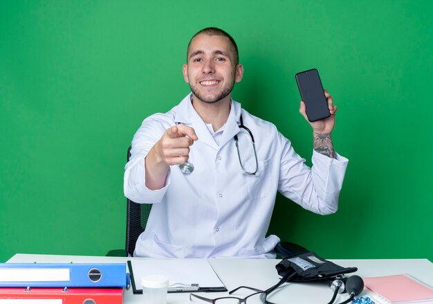 Sorridente giovane medico maschio che indossa veste medica e stetoscopio seduto alla scrivania con strumenti di lavoro che mostra il telefono cellulare e che punta davanti isolato sulla parete verde