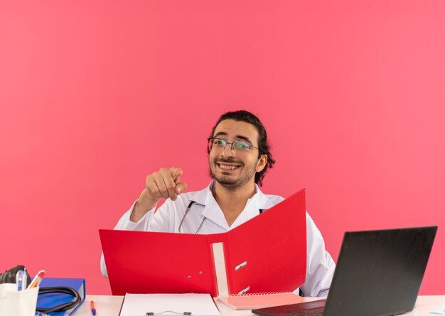 Sorridente giovane medico maschio con occhiali medici che indossano accappatoio medico con stetoscopio seduto alla scrivania