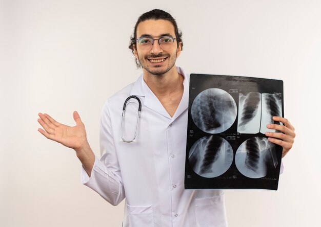 Sorridente giovane medico maschio con occhiali ottici che indossa una veste bianca con lo stetoscopio che tiene raggi x e punti con la mano a lato sulla parete bianca isolata con lo spazio della copia