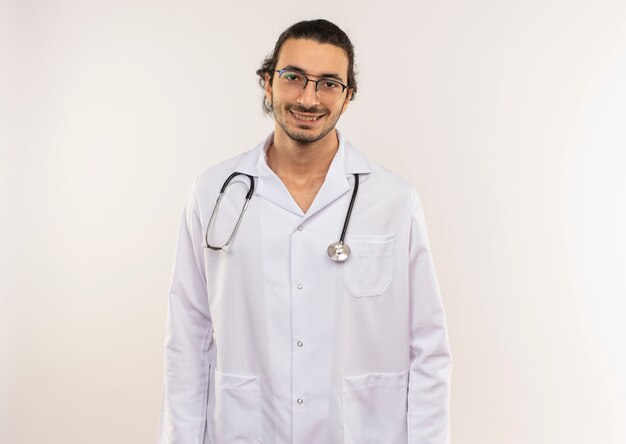 Sorridente giovane medico maschio con occhiali ottici che indossa una veste bianca con lo stetoscopio sulla parete bianca isolata con lo spazio della copia