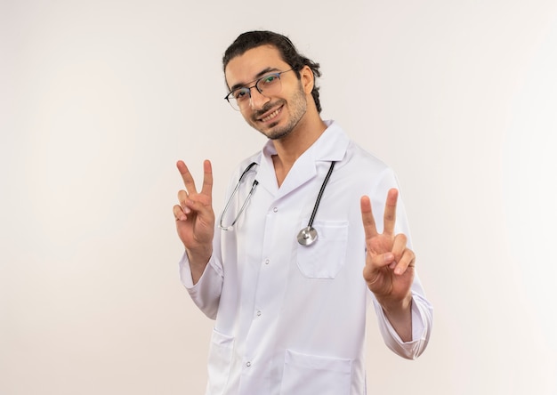 Sorridente giovane medico maschio con vetri ottici che indossa una veste bianca con lo stetoscopio che mostra il gesto di pace sulla parete bianca isolata con lo spazio della copia