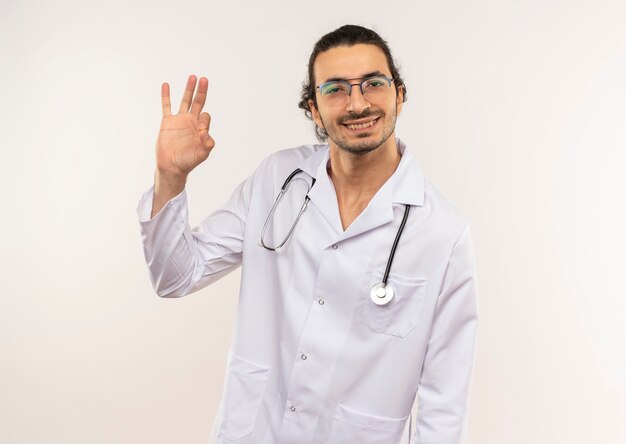 Sorridente giovane medico maschio con vetri ottici che indossa una veste bianca con lo stetoscopio che mostra okey gesto sulla parete bianca isolata con lo spazio della copia