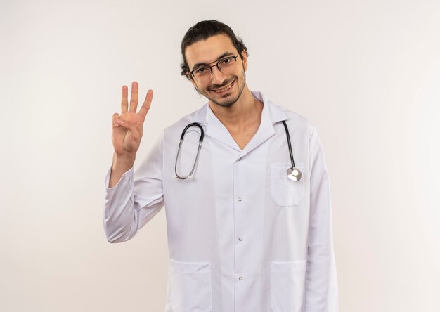 Sorridente giovane medico maschio con vetri ottici che indossa una veste bianca con uno stetoscopio che mostra tre sulla parete bianca isolata con lo spazio della copia