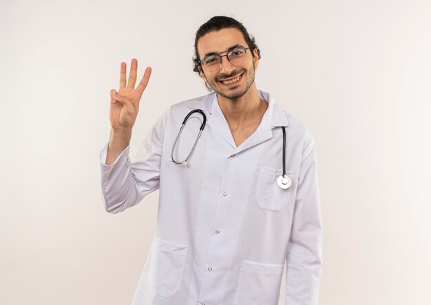 Sorridente giovane medico maschio con vetri ottici che indossa una veste bianca con uno stetoscopio che mostra tre sulla parete bianca isolata con lo spazio della copia