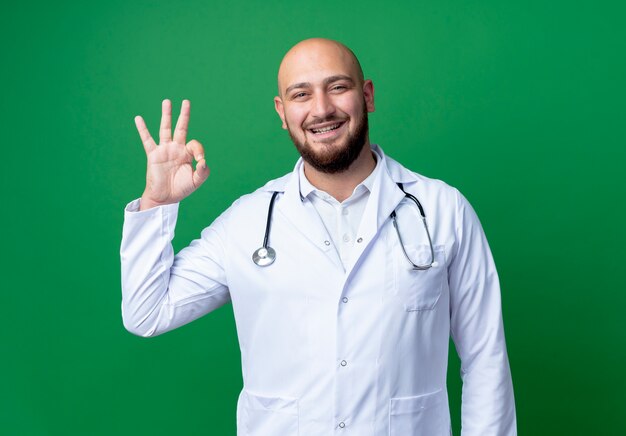 Sorridente giovane medico maschio indossa abito medico e stetoscopio che mostra okey gesto isolato su verde