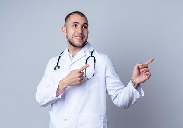 Sorridente giovane medico maschio indossa abito medico e stetoscopio intorno al collo guardando e indicando il lato isolato sul muro bianco