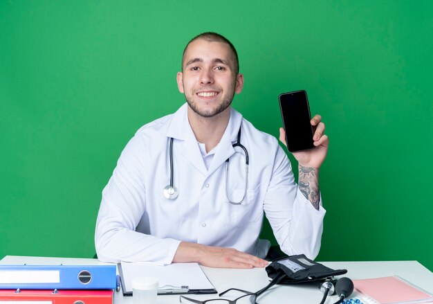 Sorridente giovane medico maschio indossa abito medico e stetoscopio seduto alla scrivania con strumenti di lavoro che mostra il telefono cellulare mettendo la mano sullo scrittorio isolato sulla parete verde