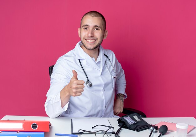 Sorridente giovane medico maschio indossa abito medico e stetoscopio seduto alla scrivania con strumenti di lavoro che mostra pollice in alto isolato sulla parete rosa