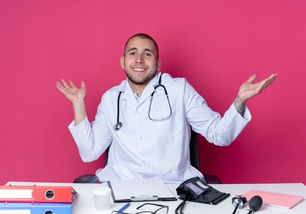 Sorridente giovane medico maschio indossa abito medico e stetoscopio seduto alla scrivania con strumenti di lavoro che mostrano le mani vuote isolate sulla parete rosa