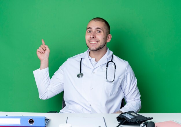 Sorridente giovane medico maschio indossa abito medico e stetoscopio seduto alla scrivania con strumenti di lavoro rivolti verso l'alto isolato sulla parete verde