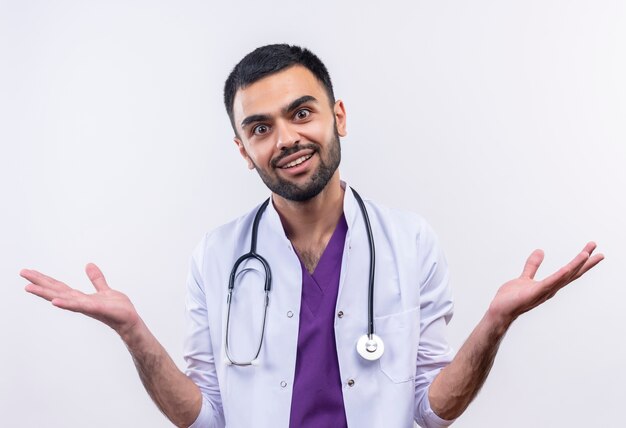 Sorridente giovane medico maschio indossa abito medico stetoscopio diffonde le mani su sfondo bianco isolato