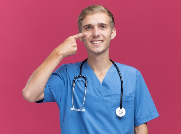 Sorridente giovane medico maschio indossa uniforme medico con lo stetoscopio tirando giù la palpebra isolata sulla parete rosa