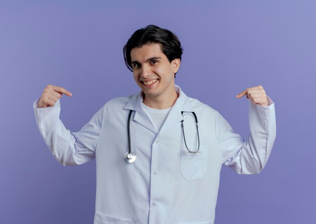 Sorridente giovane medico maschio indossa veste medica e stetoscopio e punta a se stesso isolato sulla parete viola