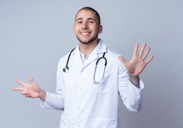 Sorridente giovane medico maschio indossa veste medica e stetoscopio intorno al collo che mostra tre e cinque con le mani isolate sul muro bianco