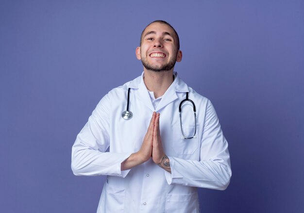 Sorridente giovane medico maschio indossa veste medica e stetoscopio intorno al collo mettendo le mani in gesto di preghiera isolato sulla parete viola