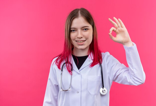 Sorridente giovane medico ragazza indossa stetoscopio abito medico che mostra okey gesto su sfondo rosa isolato
