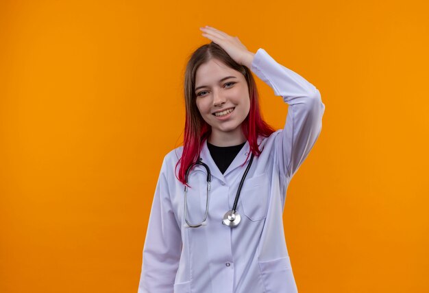 Sorridente giovane medico ragazza indossa stetoscopio abito medico mettendo la mano sulla testa isolato su sfondo arancione